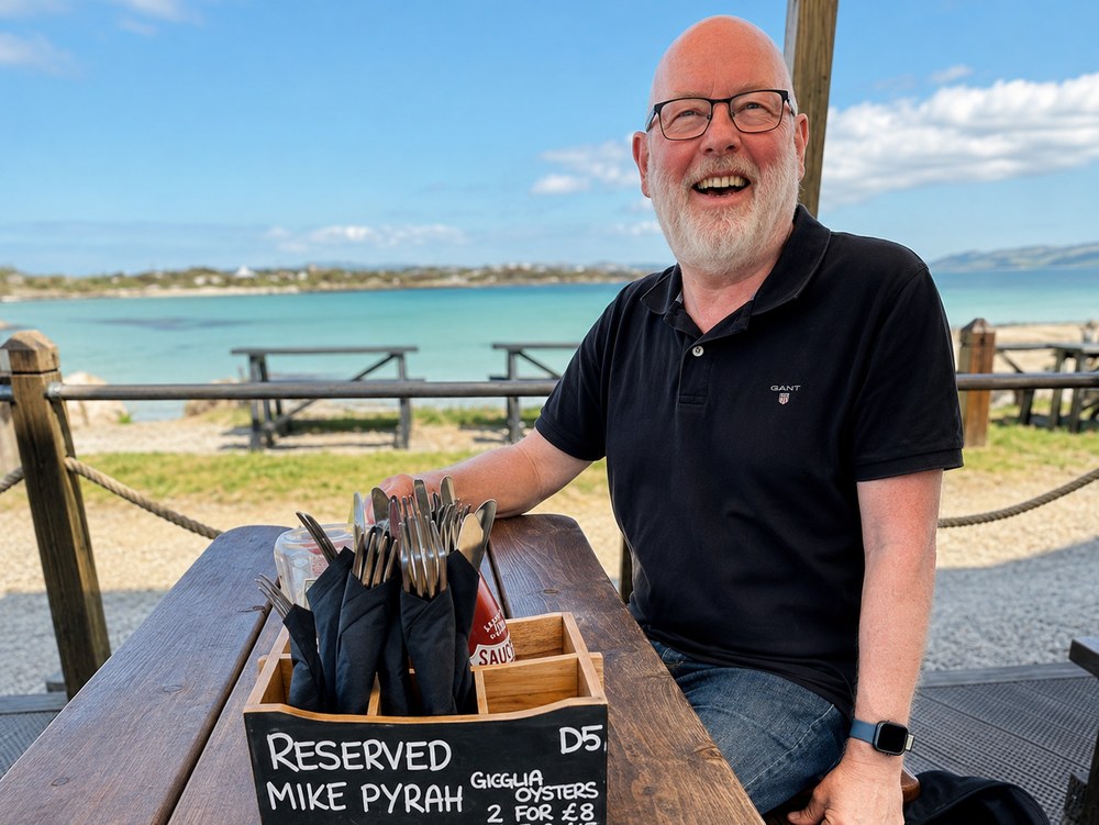 Mike Pyrah at a beachside table on Gigha, beside a chalkboard reading “Reserved Mike Pyrah”